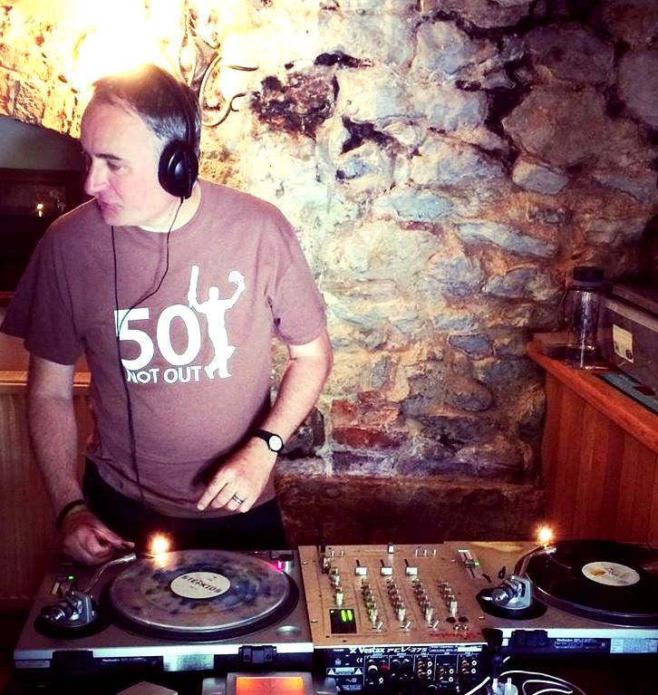 A photograph of Andy Tattersall. He is wearing headphones and appears engaged in DJing. He is wearing a T-Shirt that says '50 not out'