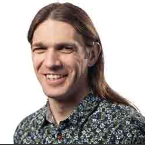 A photograph of Ben Jennings. He has long brown hair, is wearing a paisley patterned shirt and is smiling broadly
